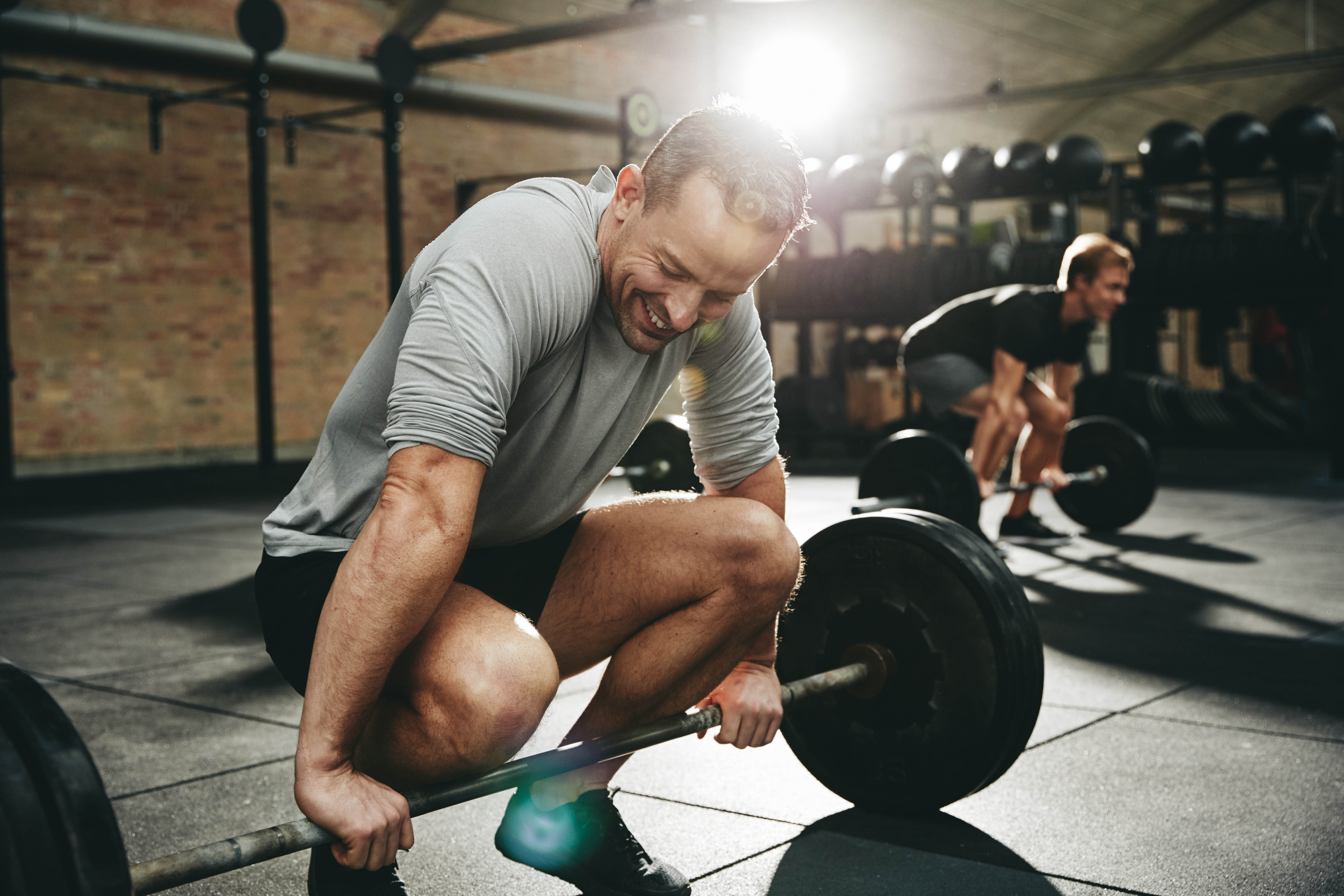 Fit man smiling while crouched down beside a barbell during a workout class at the gym Fit man smiling while crouched down beside a barbell during a workout class at the gym